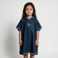 A young girl stands against a light gray studio background wearing a navy blue hooded poncho with light blue trim and a white 'Piscis' logo on the chest.